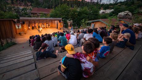 People seated in an outdoor amphitheater watching outdoor play surrounded by nature. 
