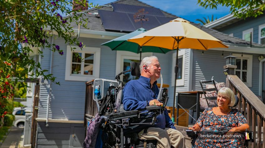 A man in wheelchair, smiling, is talking to a woman next to him in a backyard