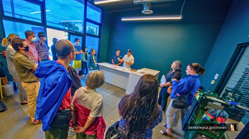 A group of people in Berkeley gather inside a modern building, listening to two presenters behind a counter during a community tour.