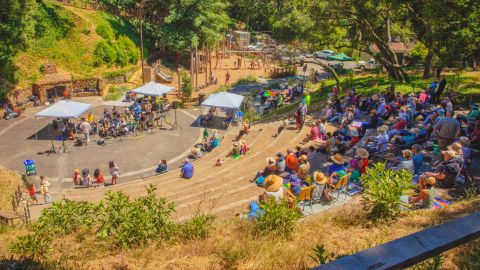A wide view of the outdoor amphitheater at Berkeley’s John Hinkel Park during a performance, with musicians playing under white tents and a large crowd seated on stone steps in the sun.