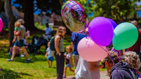 Colorful balloons, including a unicorn balloon, float above a crowd of families gathered in a Berkeley park.