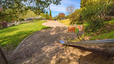 Metal slide leading to a sandy play area at Grizzly Peak Park, with open lawn, trees, and a winding path in the background.