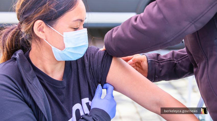A person wearing a face mask and black jacket receives a vaccination in their upper arm from a healthcare worker wearing purple gloves at a clinic in Berkeley.