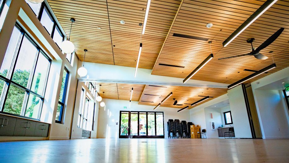 Interior of a bright, modern community room with wood-paneled ceiling, large windows, hanging lights, ceiling fans, and stacked chairs near glass doors.