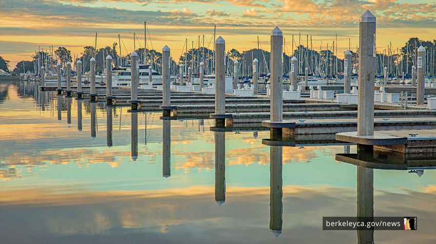 Empty marina docks and sailboat masts line calm water at sunset, with pastel sky colors reflecting across the harbor.