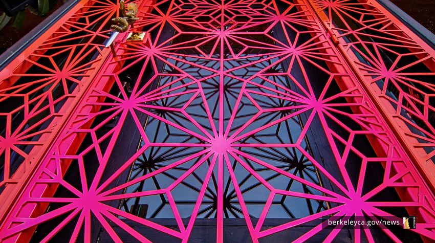 View looking up through a red geometric metal structure with a repeating lattice pattern against the sky.