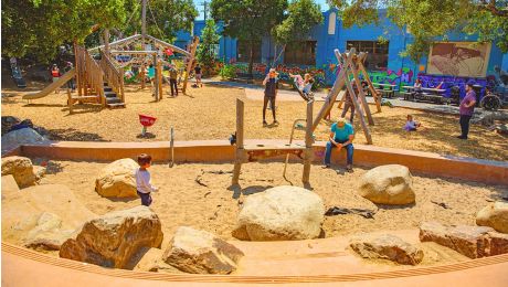 Playground with a sand play area in the foreground and a larger wood-chip play space behind it, where children climb, swing, and explore while adults sit and watch near benches and a mural-covered building.