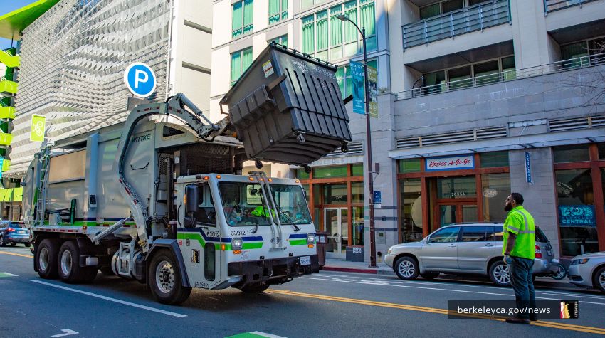 A dumpster truck lifting a dumpster at Downtown Berkeley, with a sanitation worker looking on