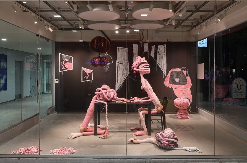 Art installation featuring two humanoid figures made of pink stuffed material sitting at a table