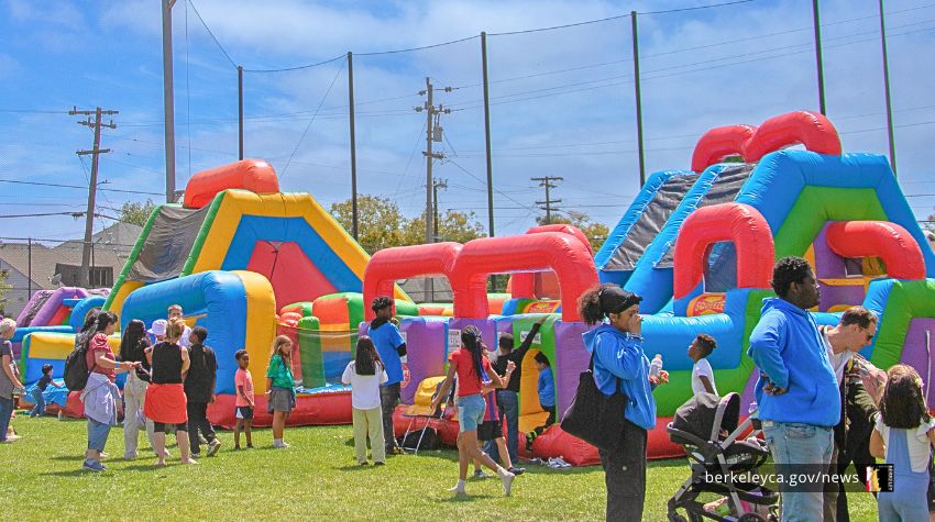 Children and families gather around colorful inflatable obstacle courses and bounce houses set up on a grassy field during a community event.