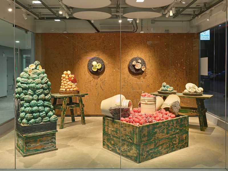 Gallery installation featuring ceramic sculptures of squash, onions, radishes, garlic, beans, and other produce displayed on weathered wooden tables, crates, and pedestals against a wood-paneled wall.