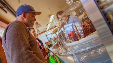 Customers stand at a bakery counter beside a glass display of baked goods.
