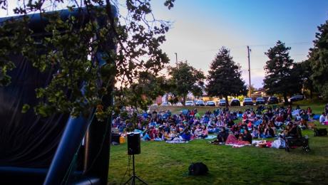 crowd sits on blankets and chairs in front of inflatable screen in a park