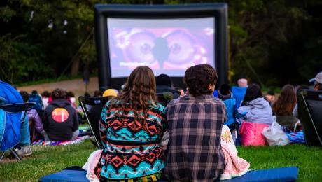 crowd sits on blankets and chairs in front of inflatable screen in a park