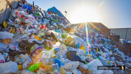 Large pile of plastic and other waste materials at a recycling facility, with sunlight shining over the top of the heap.