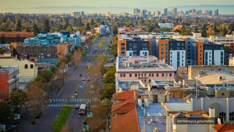 Aerial view of downtown Berkeley, showing tree-lined Shattuck Avenue with buildings on both sides and the Oakland skyline visible in the distance.