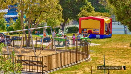 Children and families gather near a fenced-in playground with swings on the left. To the right sits a large red, yellow, and blue bounce house on the grass. Adults sit at green picnic tables under the trees, watching kids play.