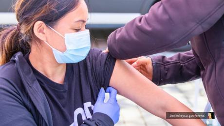 A person wearing a face mask and black jacket receives a vaccination in their upper arm from a healthcare worker wearing purple gloves at a clinic in Berkeley.