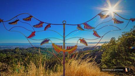 A metal sculpture stands in front of a the blue sky above the Berkeley Marina, with sunlight streaming from the top corner. White text on the image reads, “Berkeley poets: apply to become next Poet Laureate.”