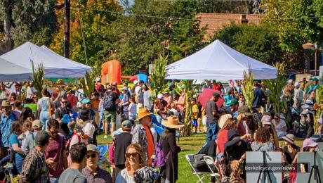 People of all ages gather on a sunny day at a lively outdoor festival with white canopy tents, hay bales, and fall decorations. Children and adults walk through the event, visit booths, and sort through a large pile of donated clothes in the foreground.