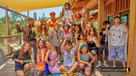 A large group of camp staff members pose together on a wooden deck outside a cabin, wearing colorful costumes and raising their hands while smiling at the camera.