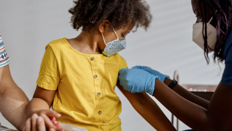 A young child receives a vaccine from a healthcare worker. The child is holding an adult's hand for comfort.