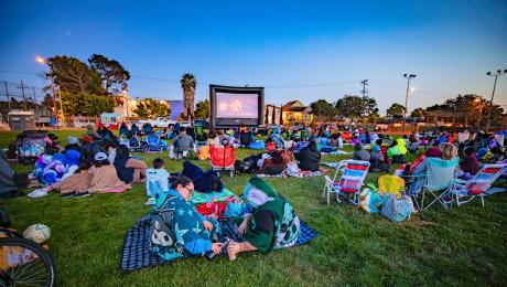 People sit on blankets and lawn chairs at dusk in a park, bundled in jackets and blankets, watching an outdoor movie on a large inflatable screen.