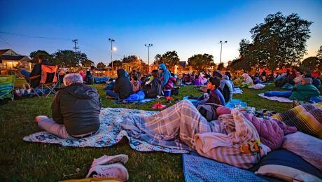 A small group sits on the grass facing a movie screen at dusk, with chairs and blankets around them.