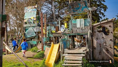 Children explore a large wooden play structure with ramps, slides, and platforms, moving through a maze of weathered boards and climbing features.