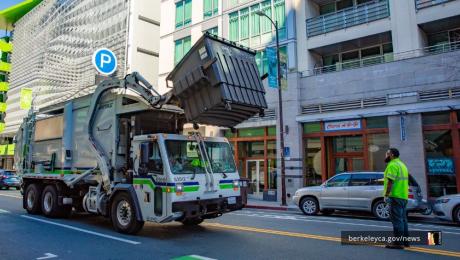 A dumpster truck lifting a dumpster at Downtown Berkeley, with a sanitation worker looking on