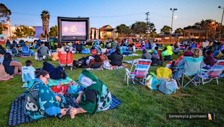 People gather on blankets and lawn chairs in a park at dusk while watching a movie on a large outdoor screen.
