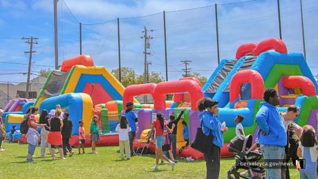 Children and families gather around colorful inflatable obstacle courses and bounce houses set up on a grassy field during a community event.