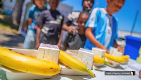 Bananas and milk cartons sit on a table in the foreground while children gather around.