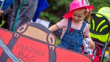 A child behind a cardboard cutout of an emergency bag looking excited.