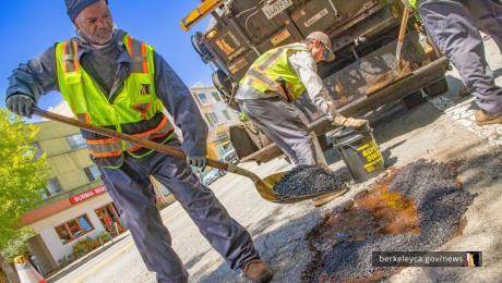 Public Works staff shovels black asphalt in to a hole, during pothole repair. 