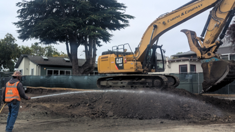 The soil remediation in progress. An excavator sits on top of excavated dirt. A worker is spraying the mound of dirt to reduce dust in the air.